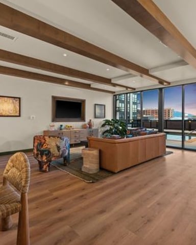 Modern loft living room with exposed wood beams, brown leather sofa, and floor-to-ceiling windows overlooking city skyline at sunset