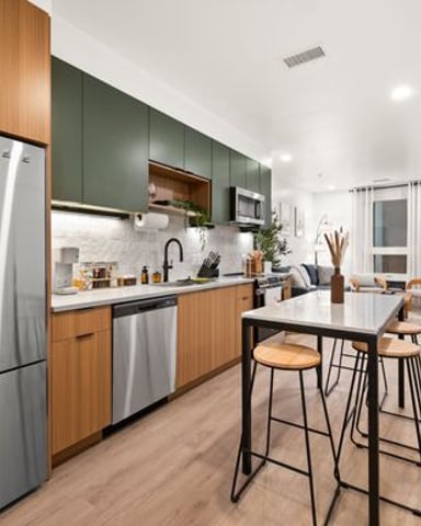Modern kitchen with wood cabinetry, stainless steel appliances, green upper cabinets, and white quartz countertops, opening to dining area with bar seating