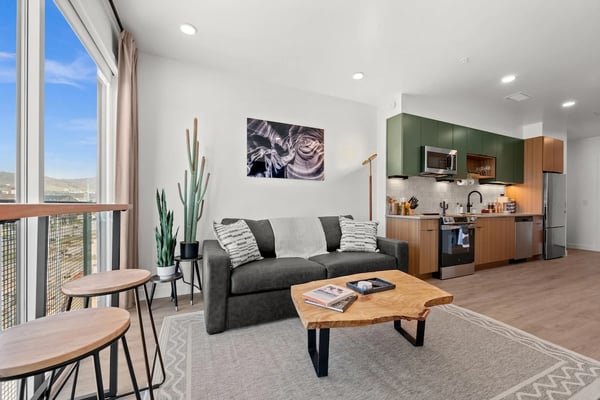 Open-plan living room with gray sofa, wooden coffee table, kitchen with green cabinets, and mountain view from balcony